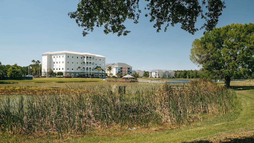 A large white building with multiple floors and balconies, surrounded by a grassy area with a pond and reeds.