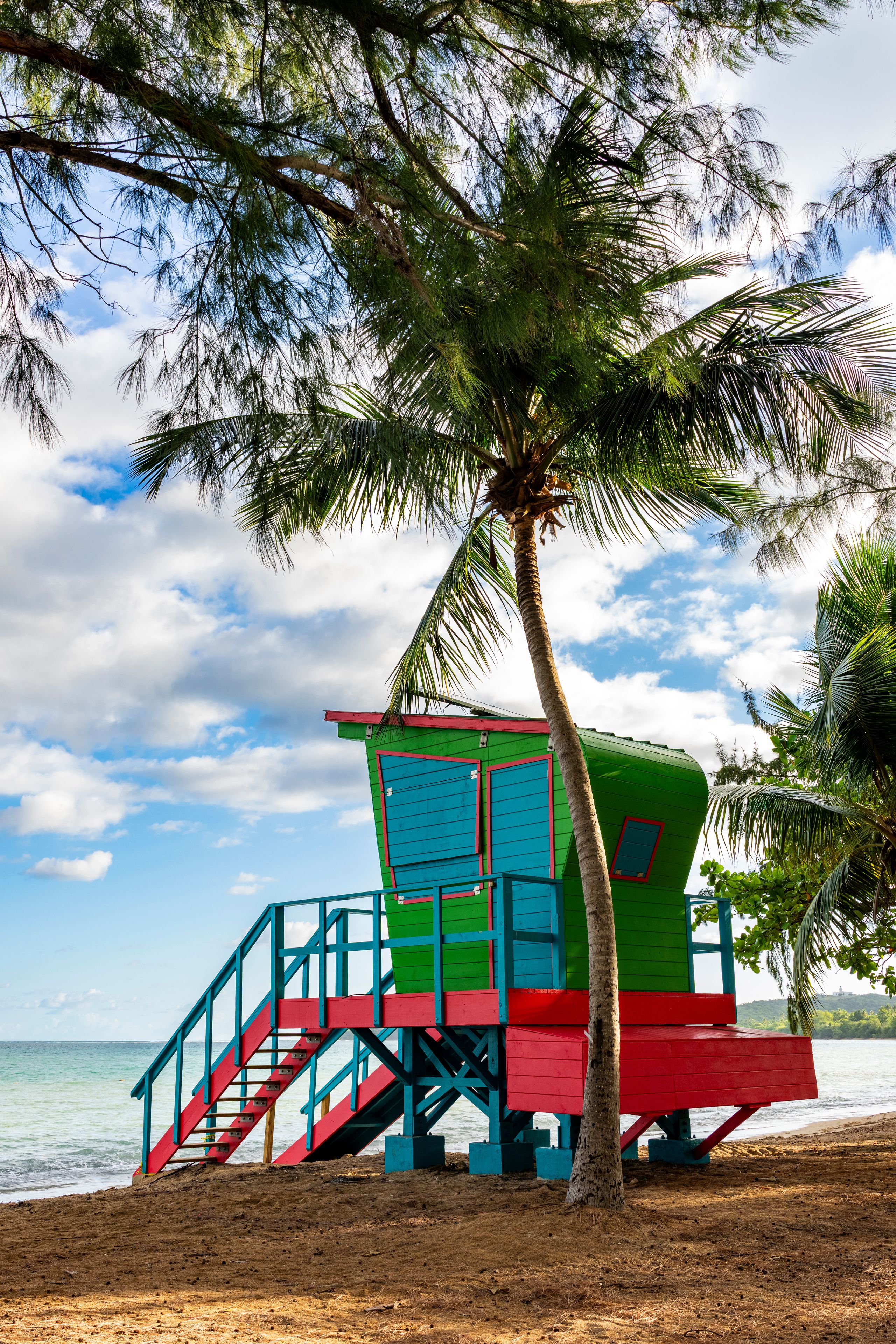 Uma torre colorida de salva-vidas com escadas, uma palmeira e uma praia com o oceano ao fundo.