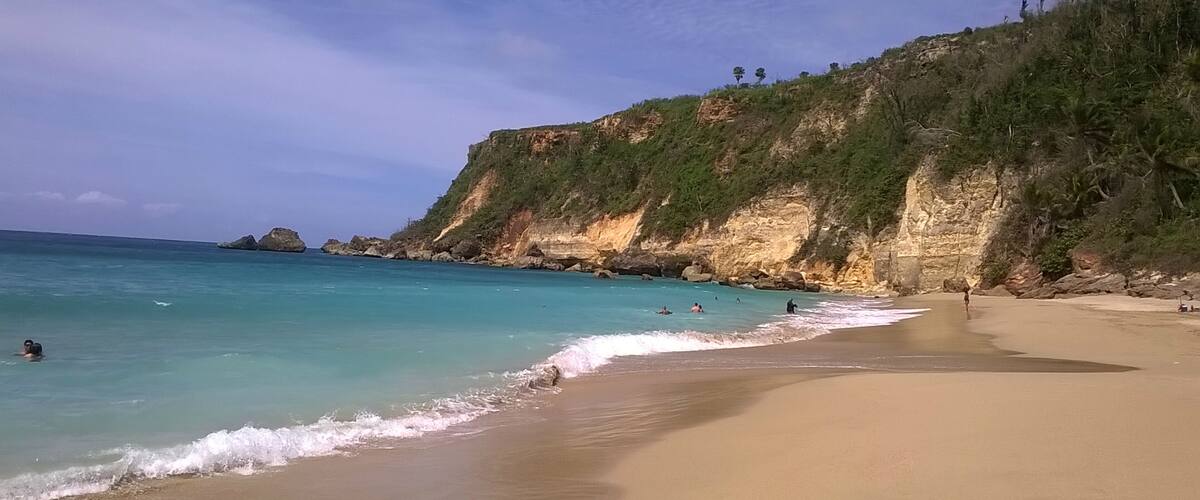 A beach with clear turquoise water, a sandy shore, and rocky cliffs with sparse vegetation.