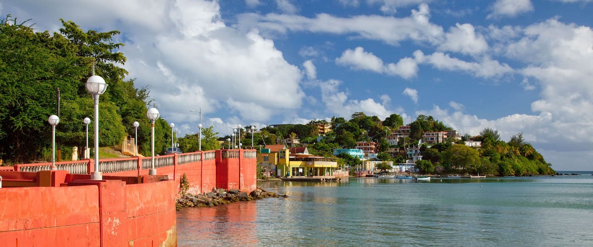 A coastal scene with a red barrier, clear blue water, and buildings on a hillside.