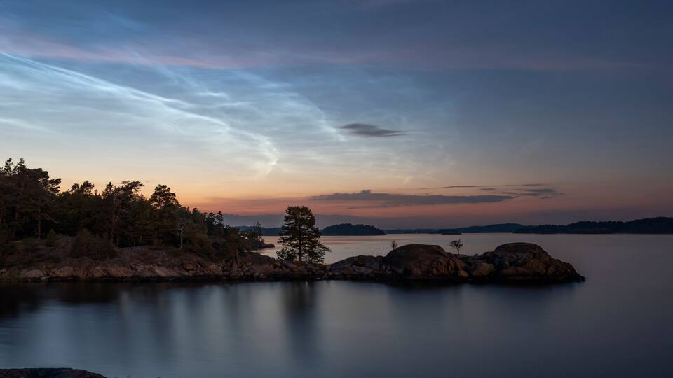 A calm lake at sunset with a rocky shoreline and trees.