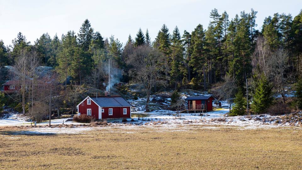 A rural landscape with a red barn, a wooden cabin, and a forested area with snow patches.