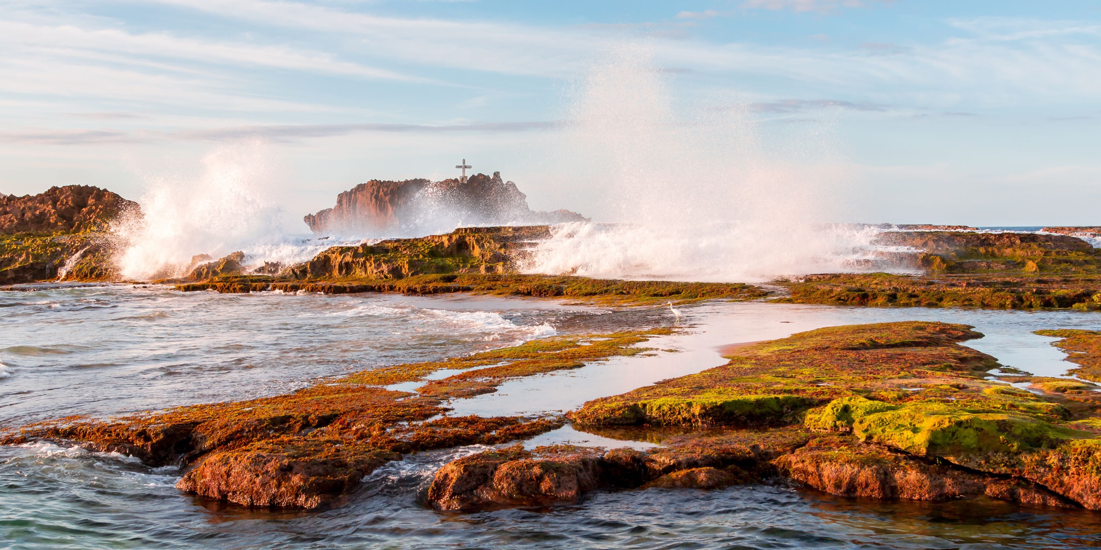 A rocky coastline with waves crashing against the rocks and a cross on top of a cliff.