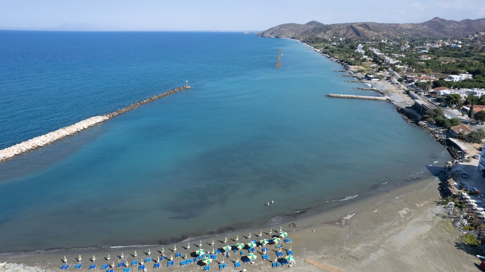 A coastal town with a clear blue sea, a rocky breakwater, and a row of beach umbrellas.