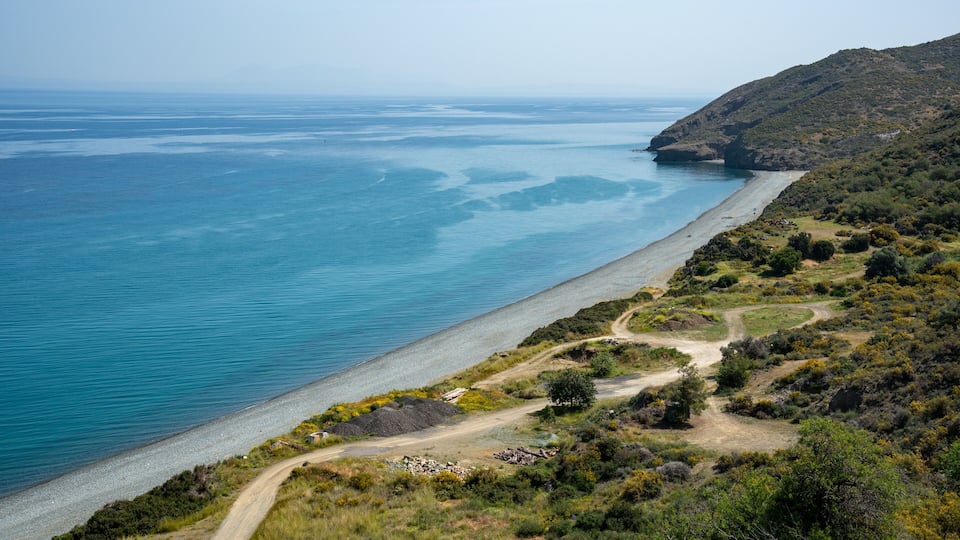 A coastal landscape with a clear blue sea, a sandy beach, and a dirt path winding through the hills.