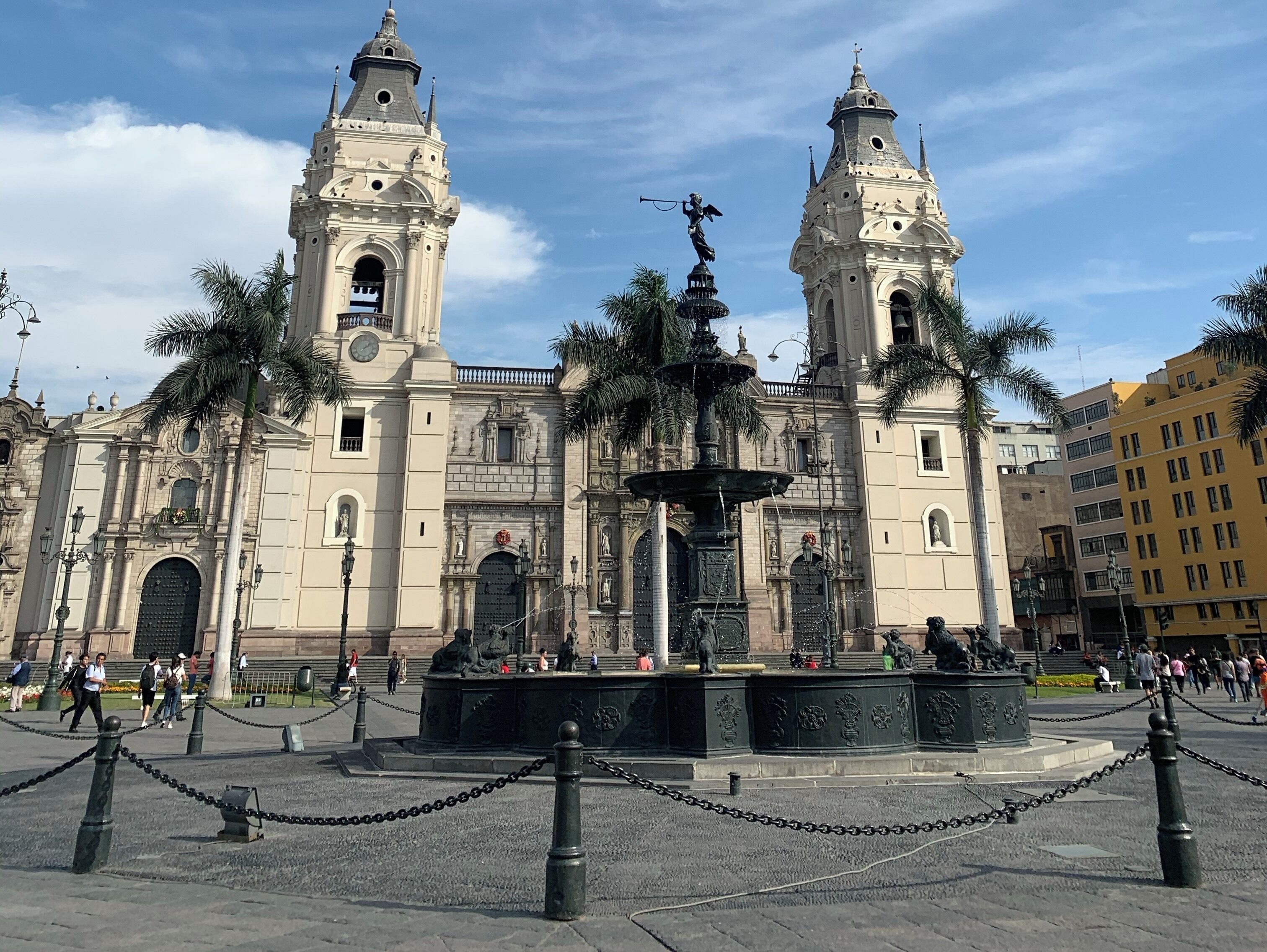 Una iglesia histórica con dos torres, una fuente y palmeras al frente.