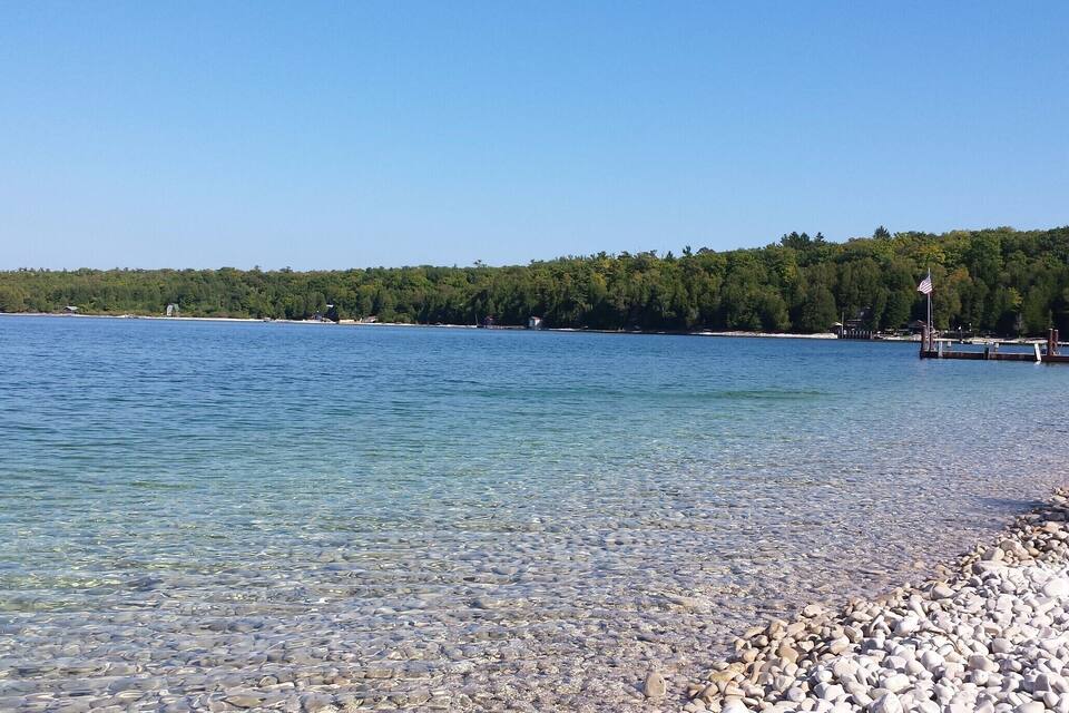 Een helder meer met een kiezelstrand en een bebost heuvel in de verte.