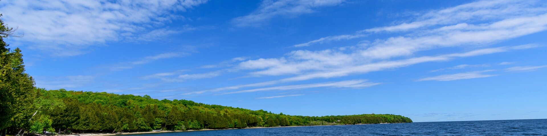 Una playa de guijarros con agua cristalina, una isla cubierta de bosque y un cielo parcialmente nublado.