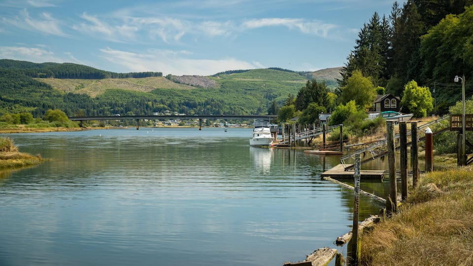 A calm river with a boat docked on the right, a bridge in the background, and a forested hillside.