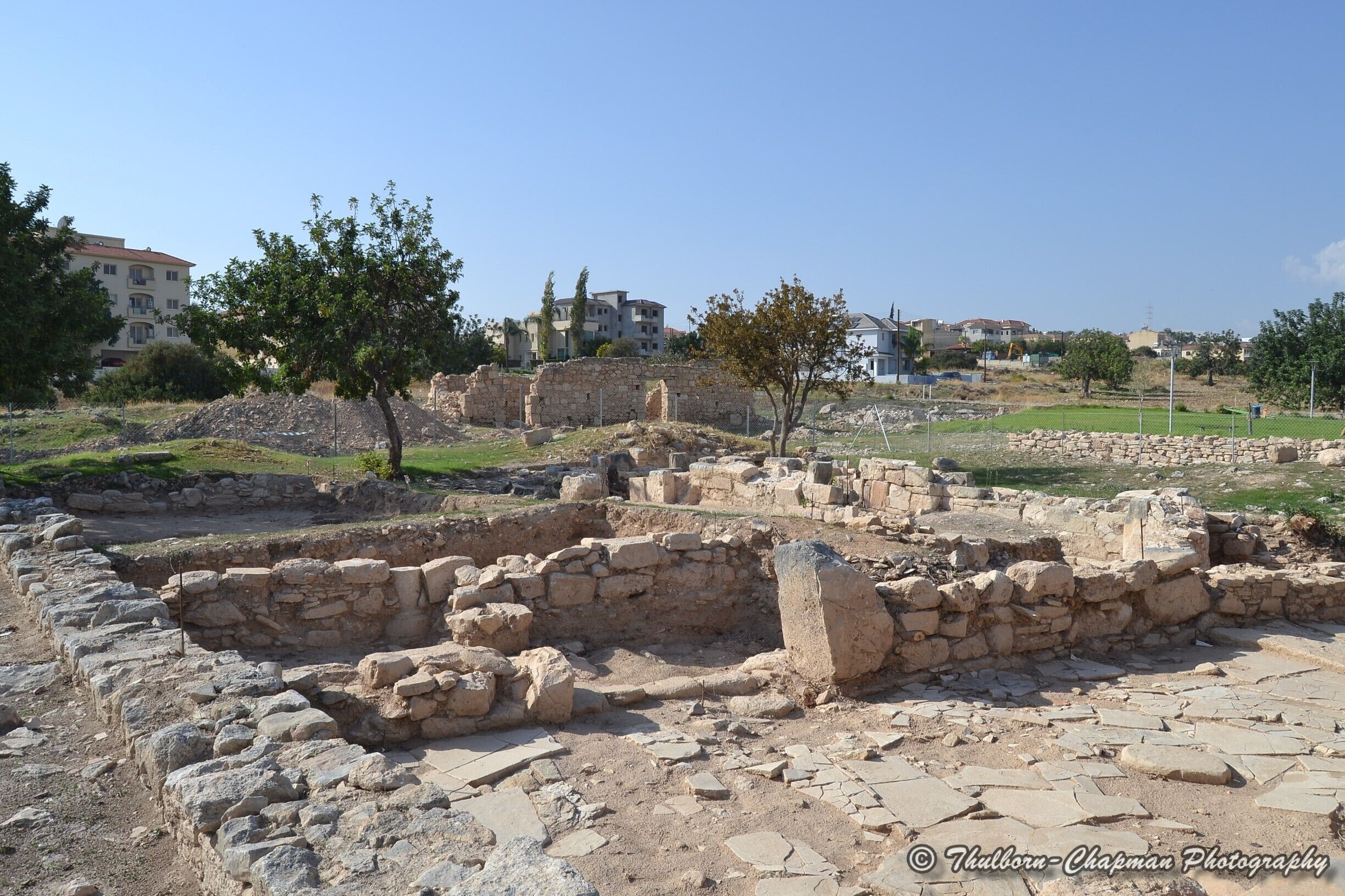 Ancient ruins with stone walls and scattered stones, surrounded by trees and modern buildings in the background.