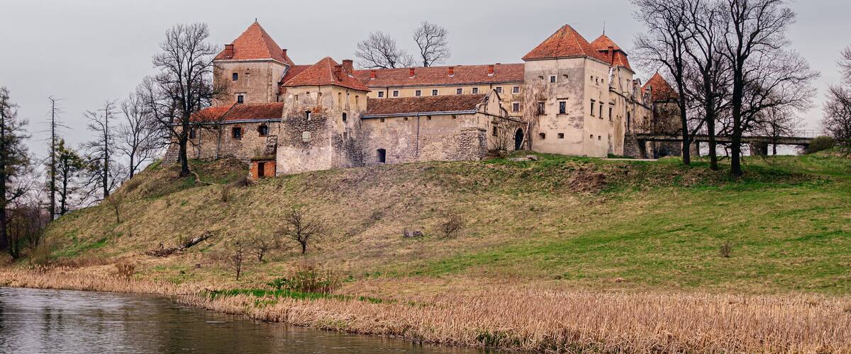 A historic castle with red-tiled roofs, situated on a grassy hill by a calm river.