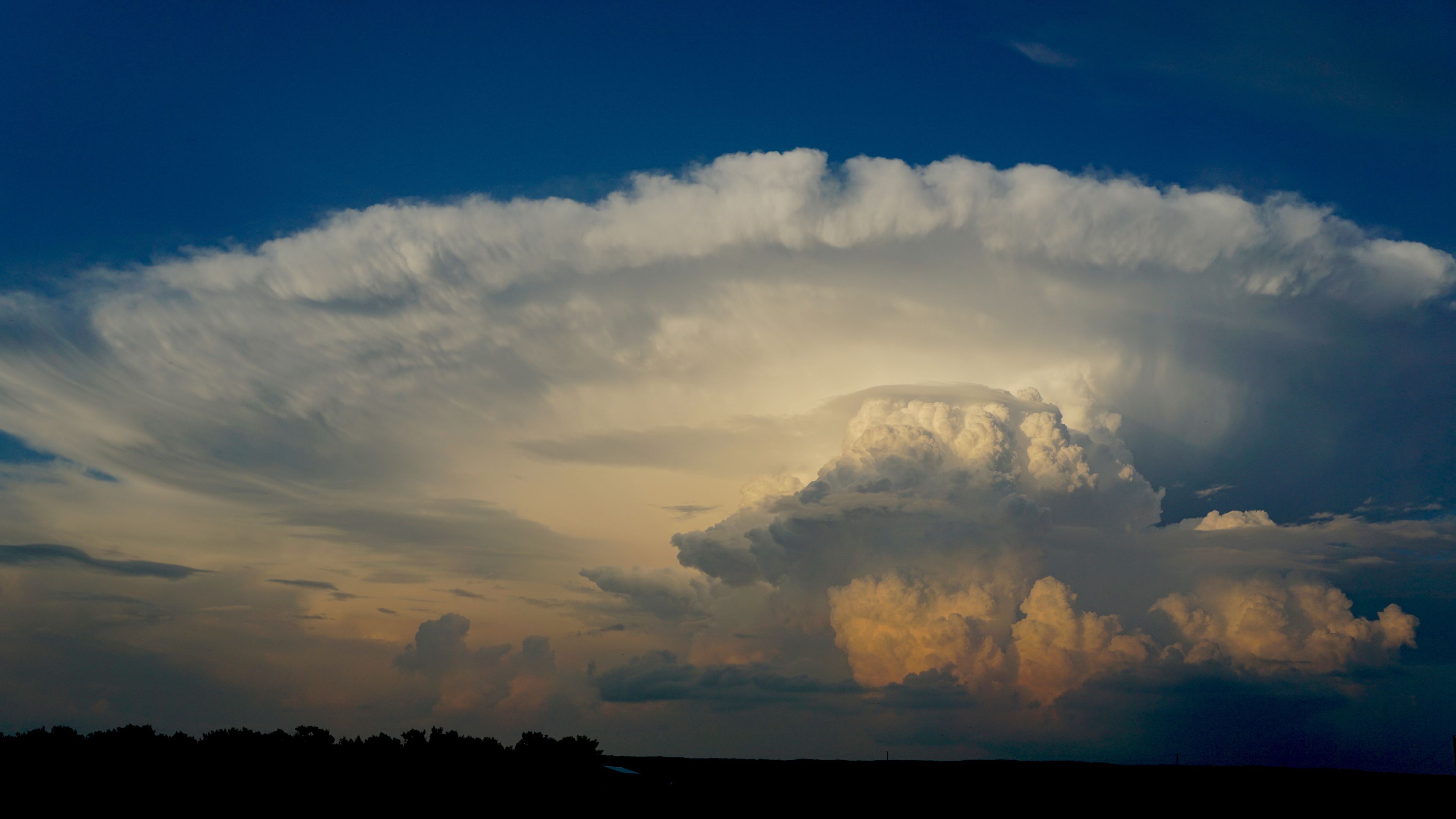 A dramatic sky with a large cloud formation and a silhouette of a tree line at the horizon.