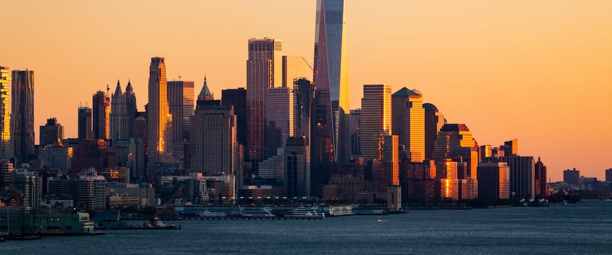 A city skyline at sunset with prominent skyscrapers and a body of water in the foreground.
