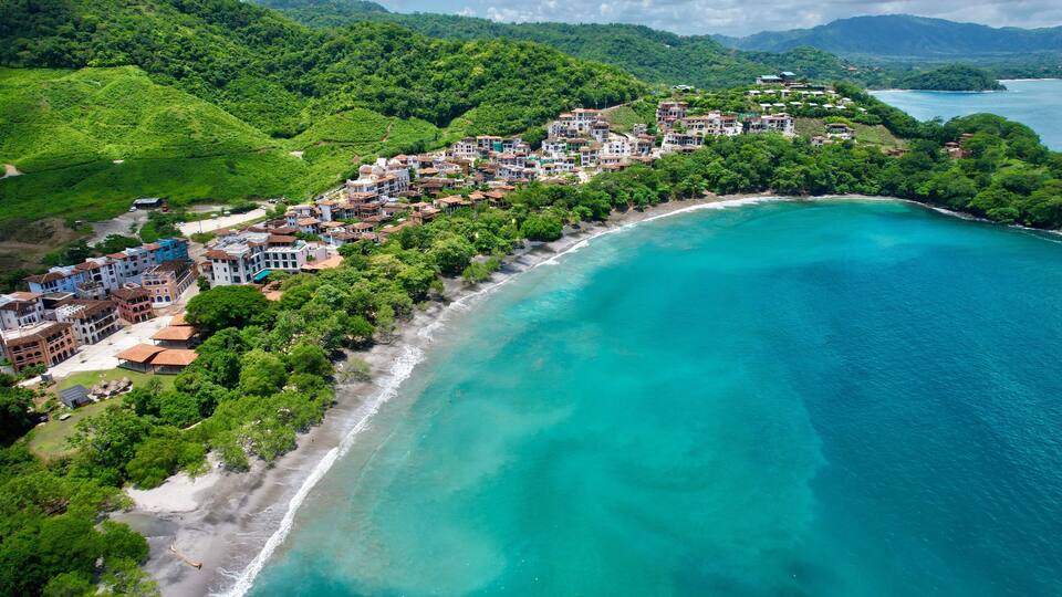A coastal town with buildings along a turquoise bay, surrounded by lush green hills.
