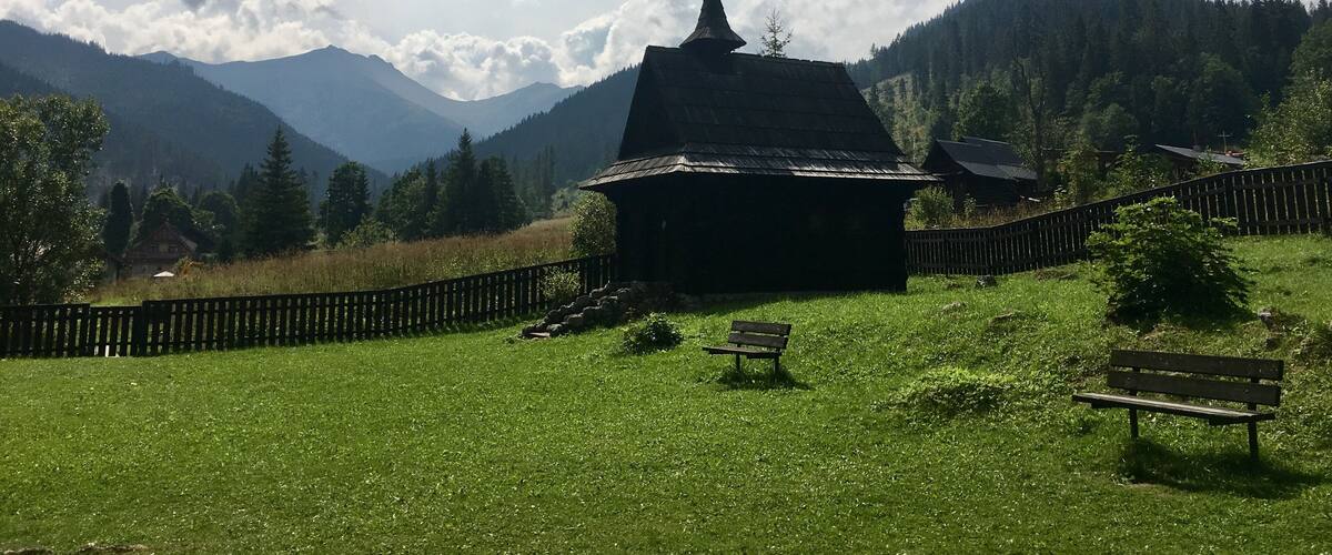 A wooden house with a pointed roof surrounded by a wooden fence, set against a backdrop of mountains and a partly cloudy sky.