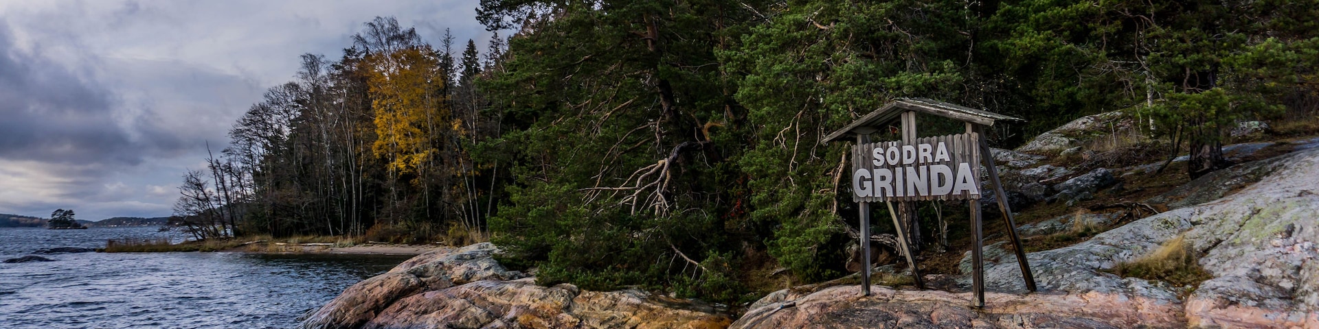 Una costa rocosa con un letrero de madera que dice 'SODRA GRINDA' en medio de un bosque frondoso y una masa de agua.