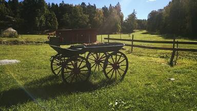 A wooden cart in a grassy field with a wooden fence and a red barn in the background.