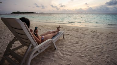 Back view of unrecognizable female tourist in bikini sitting on chair and browsing mobile phone while spending evening on sandy beach during summer holidays on Maldives islands