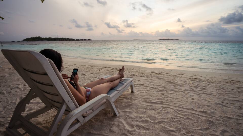 A person relaxing on a beach chair, looking at a smartphone.