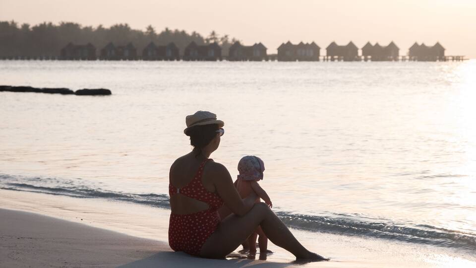 A woman and a child sitting on a beach with a row of houses in the background.