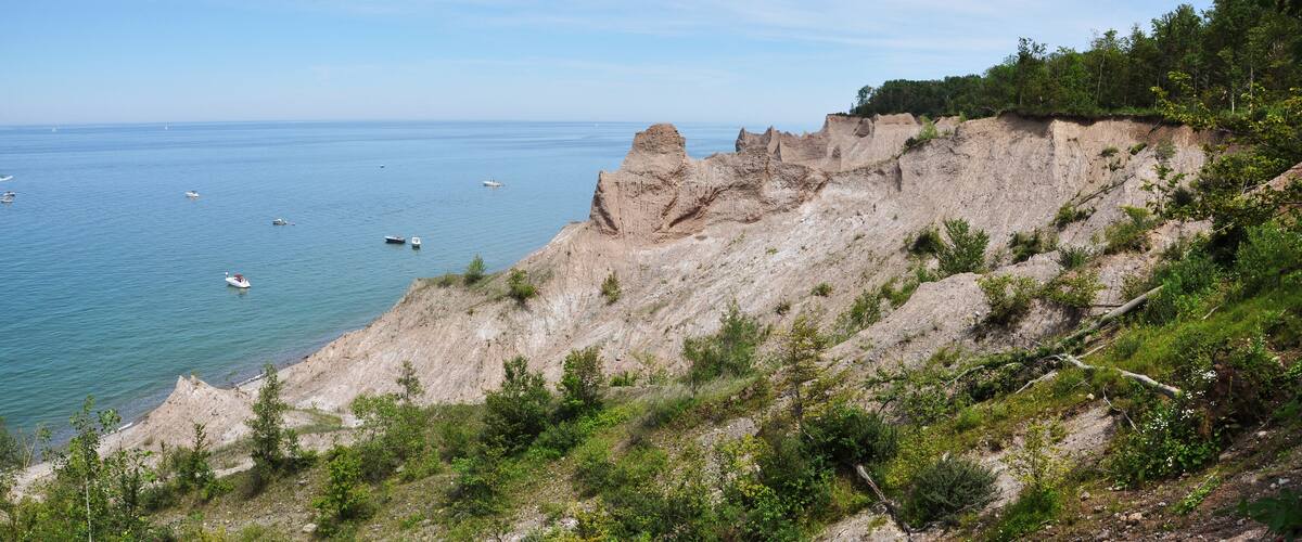 A coastal cliff with sparse vegetation and a calm sea with several boats.