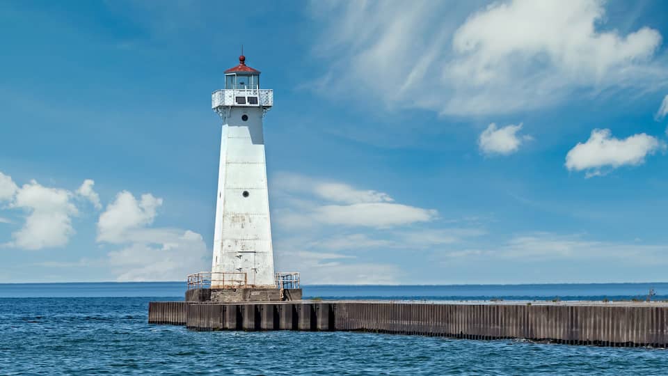 A white lighthouse on a wooden pier extending into the ocean under a partly cloudy sky.