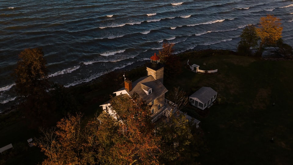 A coastal house with a lighthouse, surrounded by trees and ocean waves.