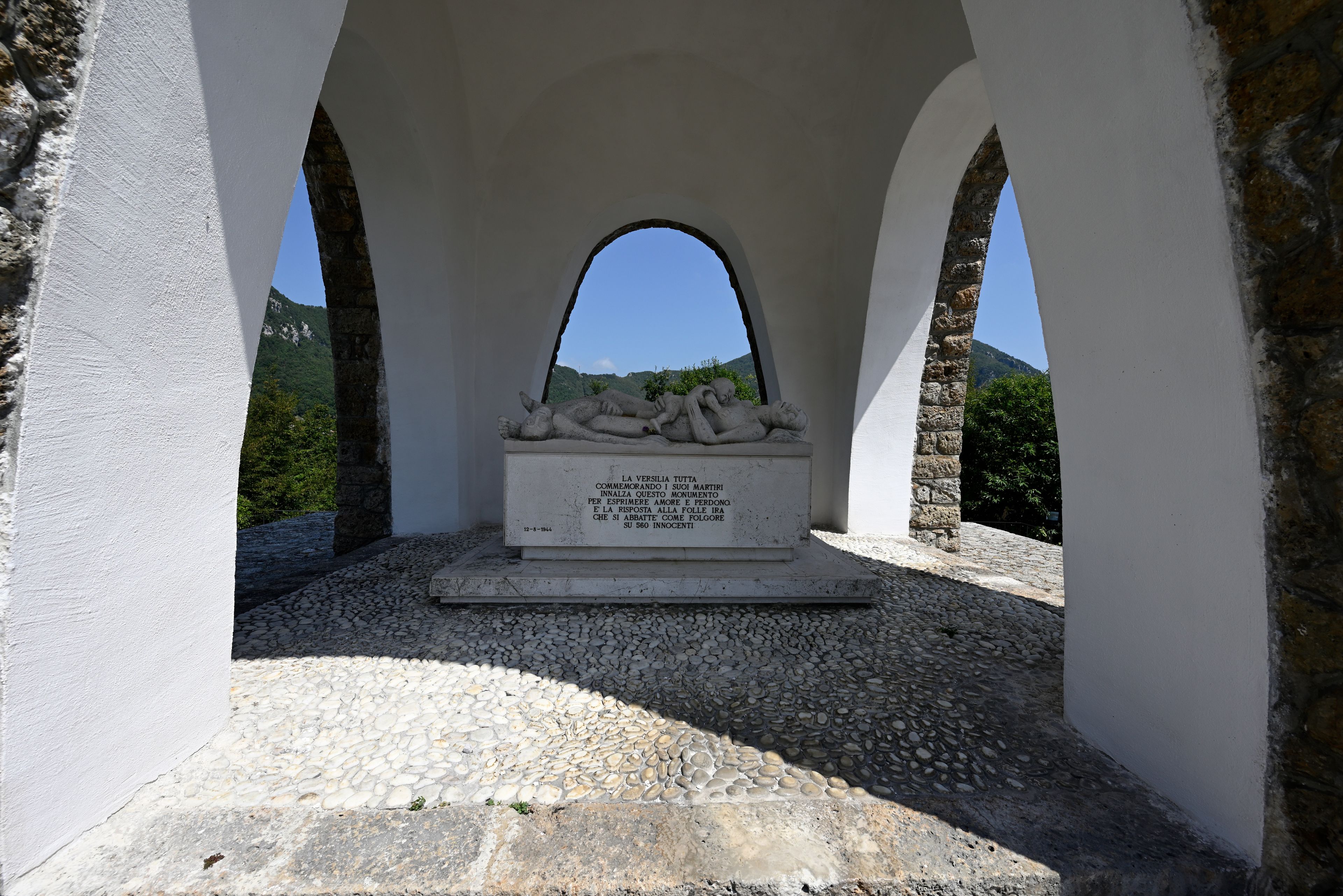 A memorial with a stone sculpture of two figures under an arched structure, surrounded by white walls and a clear sky.
