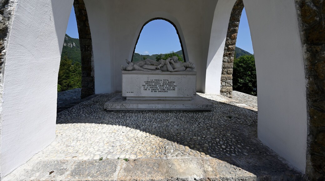 A memorial with a stone sculpture of two figures under an arched structure, surrounded by white walls and a clear sky.