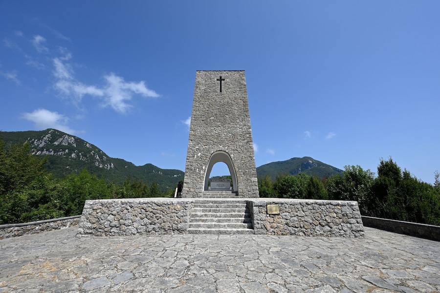 Um monumento de pedra com uma cruz no topo, rodeado por um muro de pedra e degraus que levam até ele, com montanhas ao fundo.