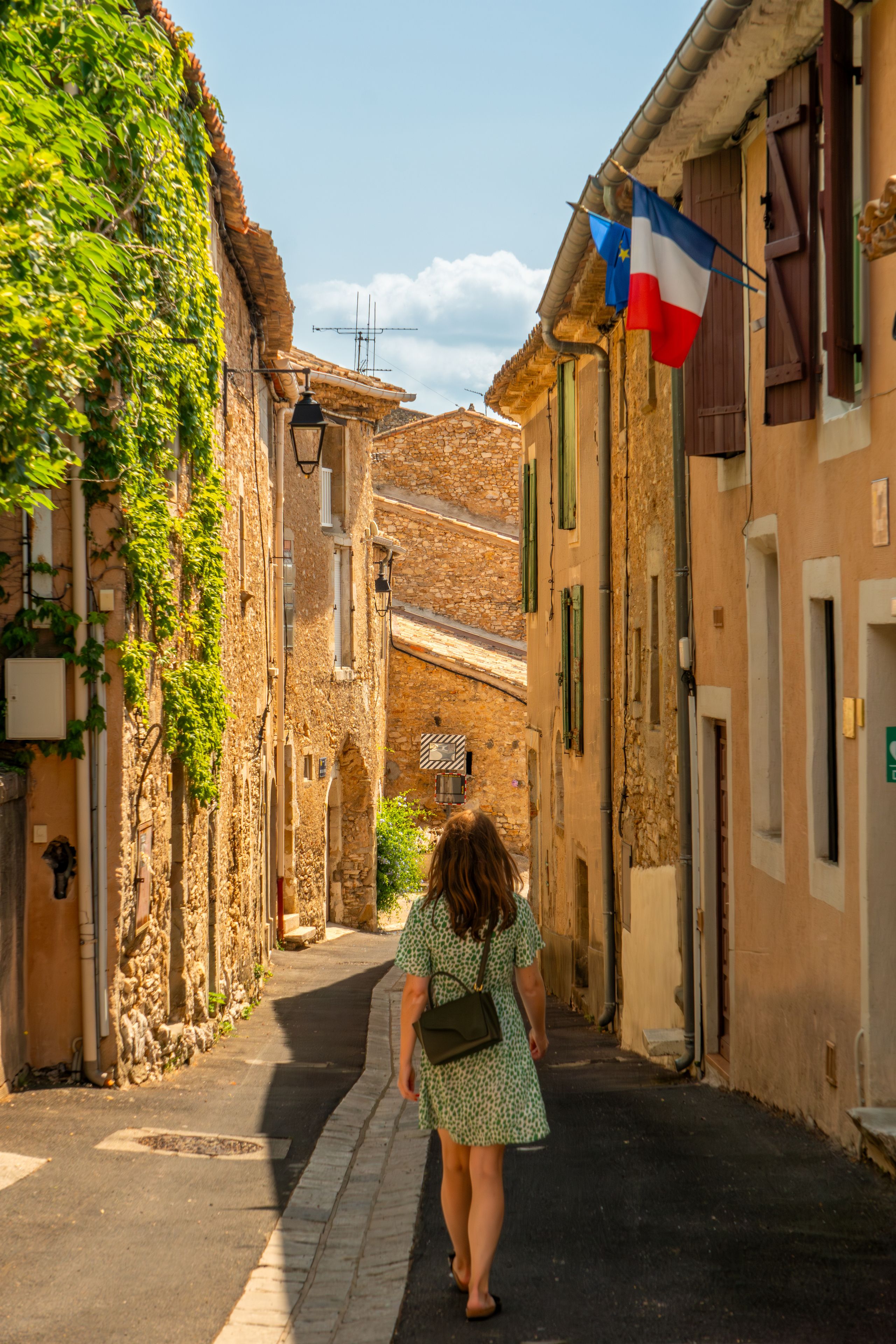 A narrow street with stone buildings, a woman walking, and flags.