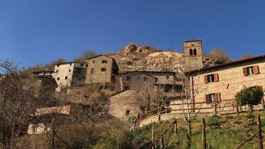 A stone village with buildings on a hillside, a bell tower, and a wooden bridge.