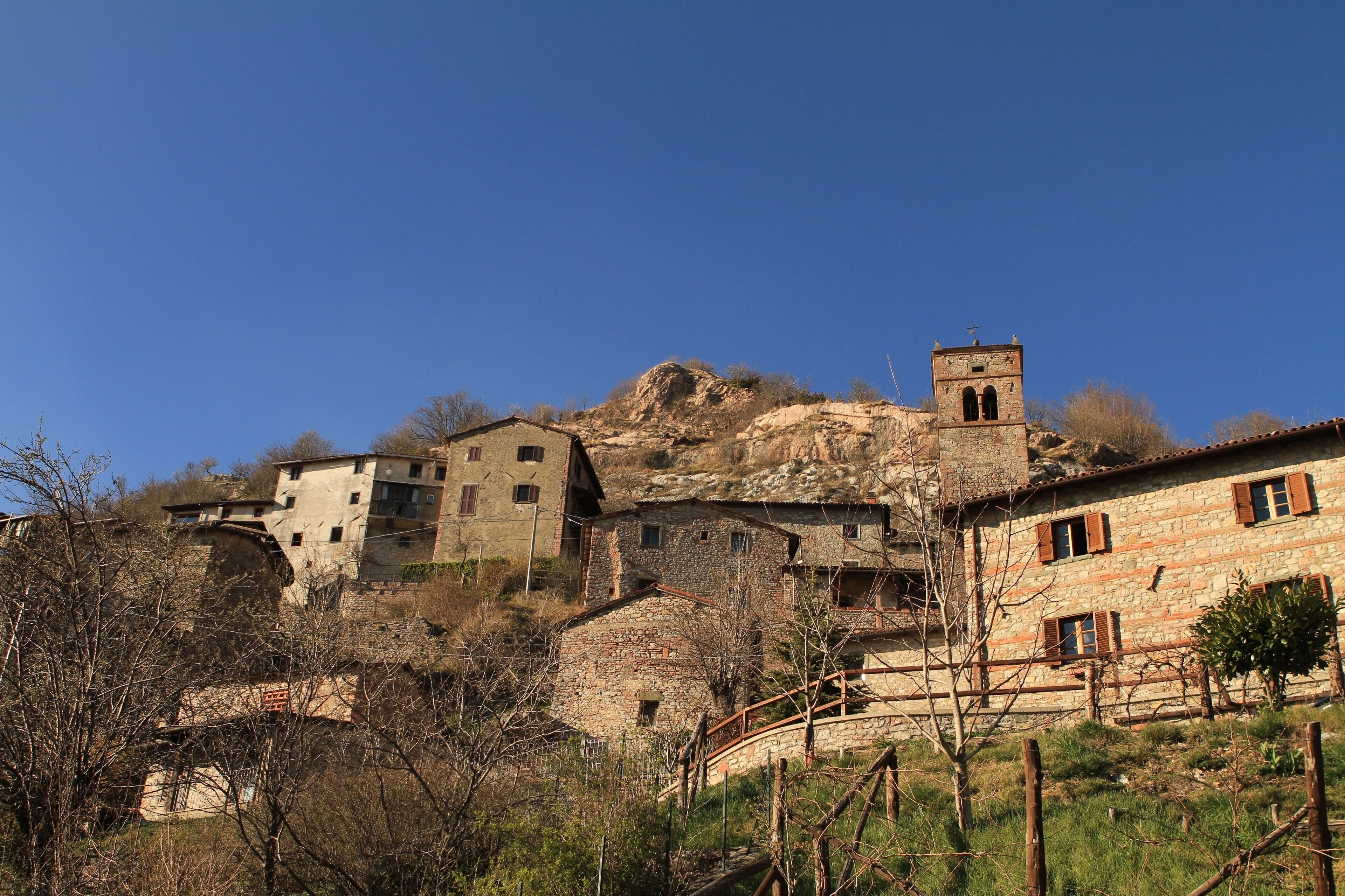 A stone village with buildings on a hillside, a bell tower, and a wooden bridge.