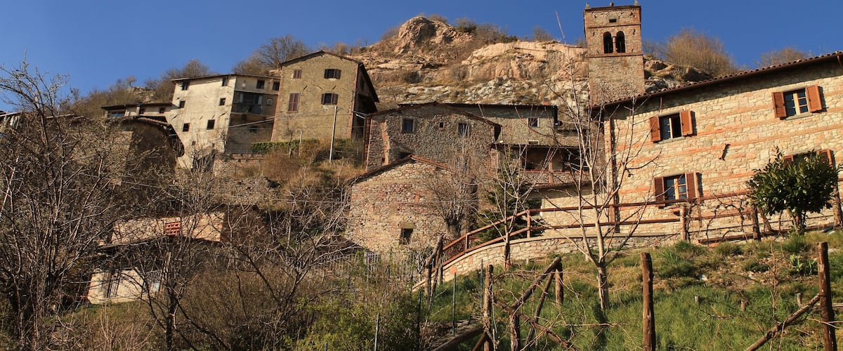A stone village with buildings on a hillside, a bell tower, and a wooden bridge.