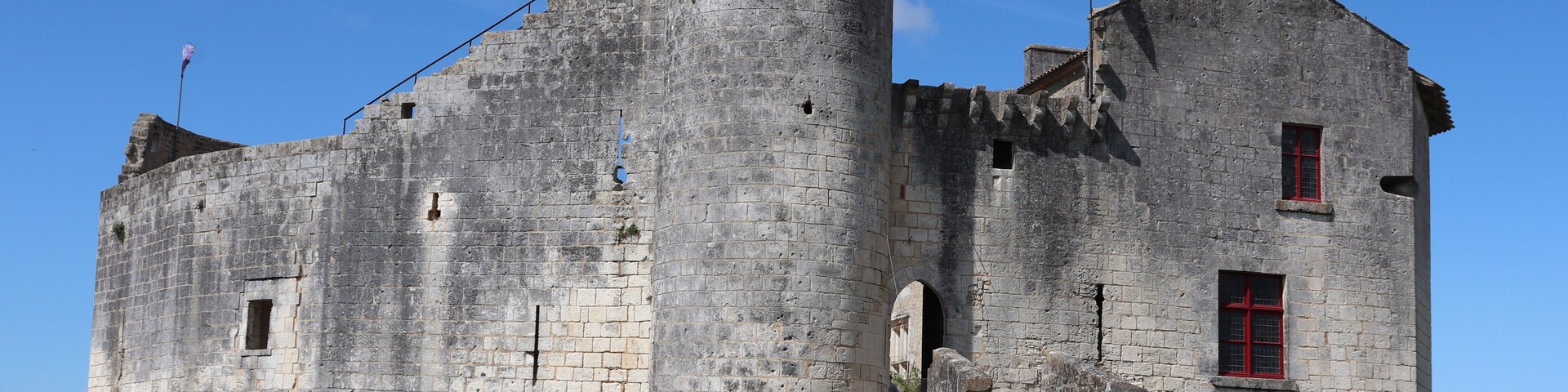 A historic stone castle with a flag on top, a red window, and a clear blue sky.