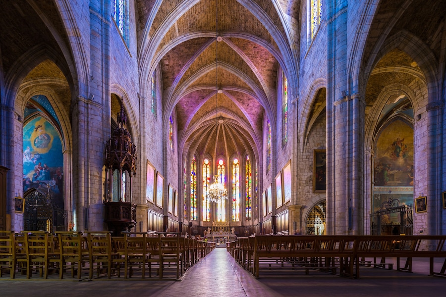 Interior de una catedral con altos techos abovedados, vidrieras de colores y filas de bancas de madera.
