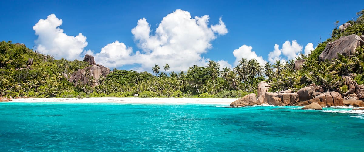 A turquoise beach with white sand, palm trees, and large rock formations under a clear blue sky.