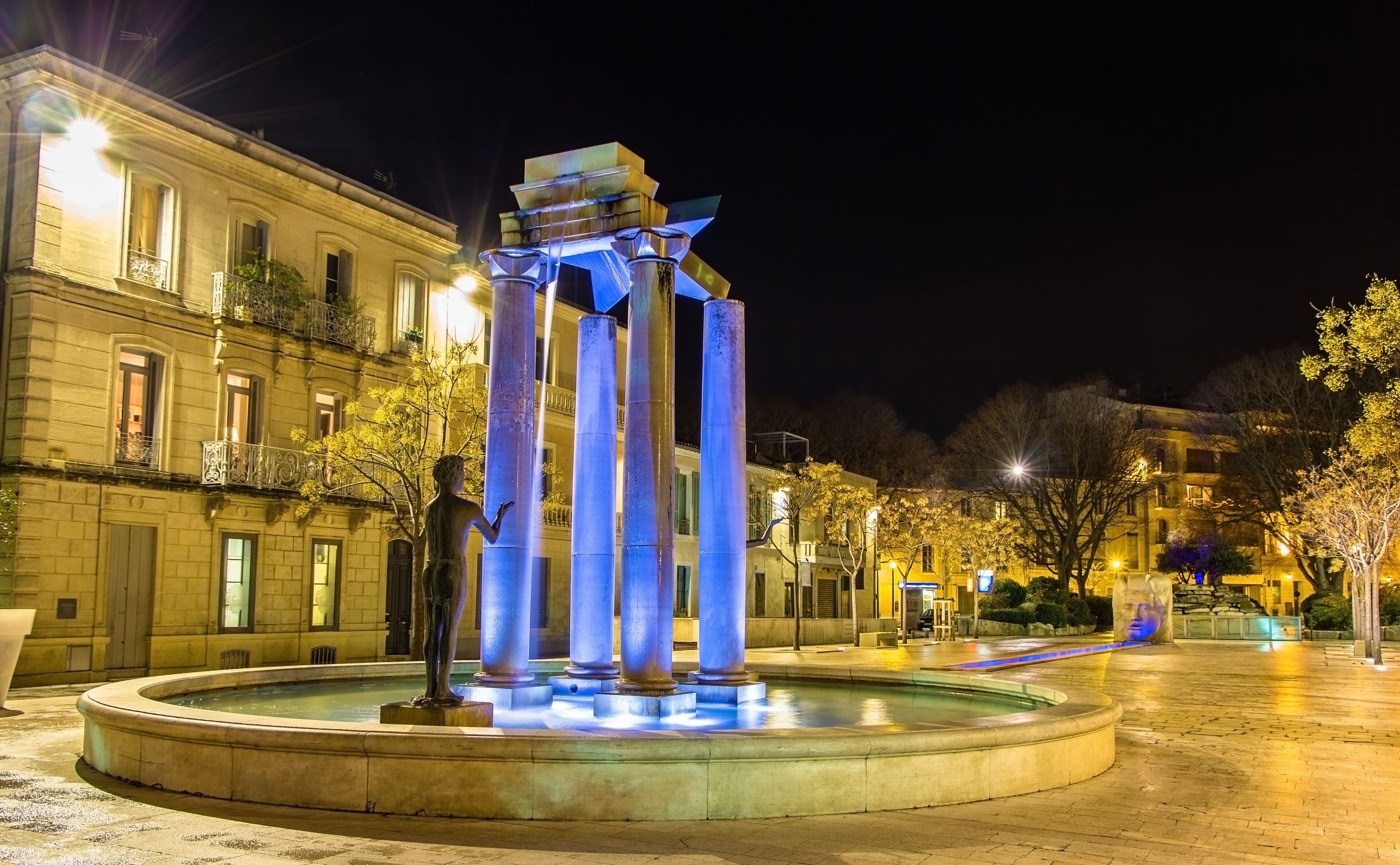 A night scene with a fountain featuring a statue and illuminated columns.