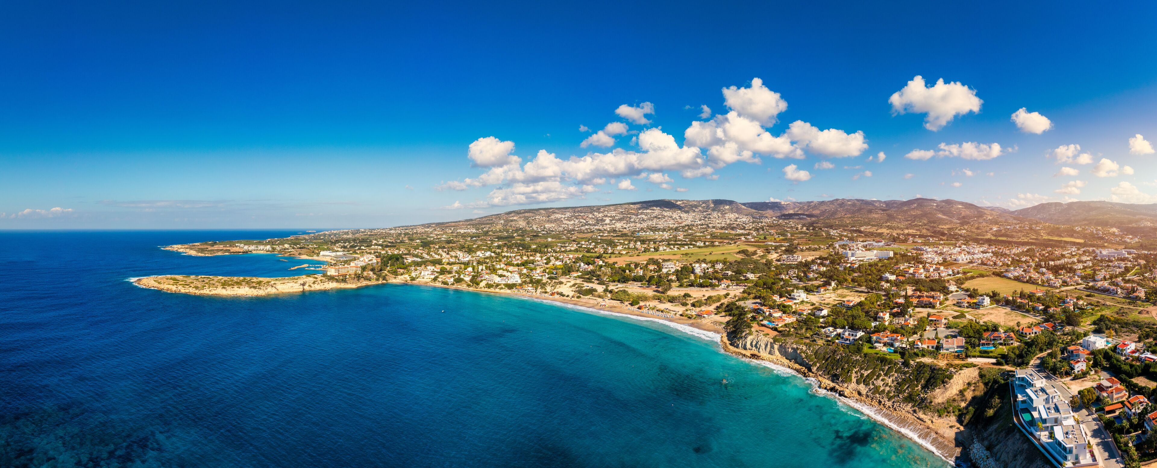 A coastal town with a beach, clear blue waters, and a mountainous backdrop.