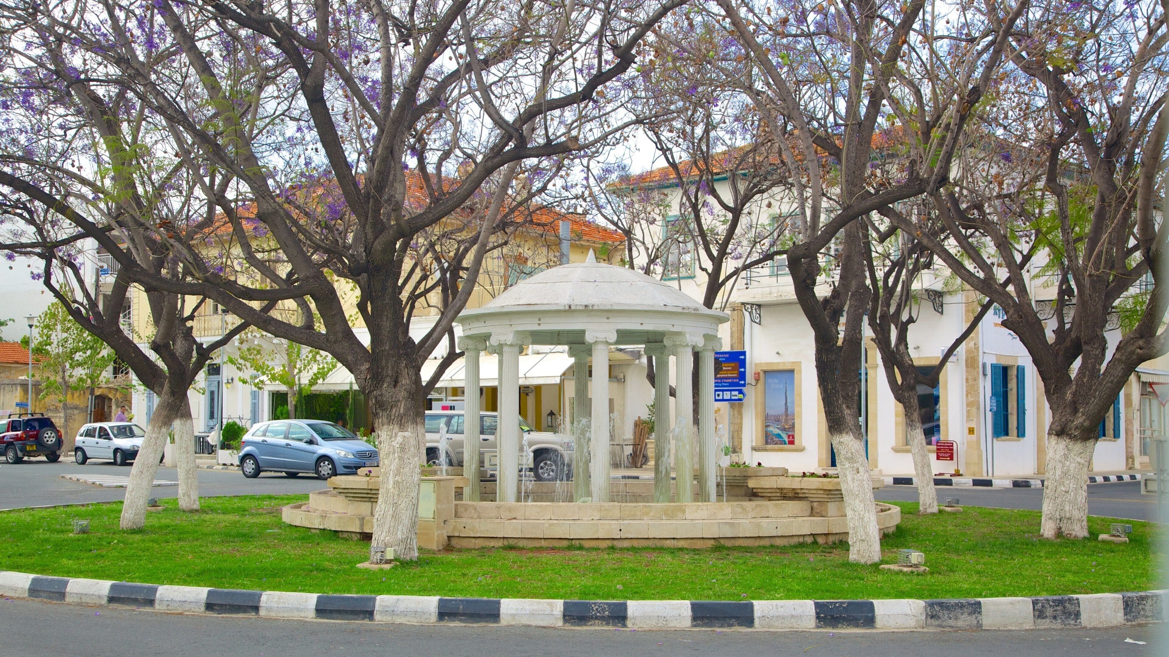 A gazebo surrounded by trees with purple flowers, a white building with blue shutters, and parked cars on a street.