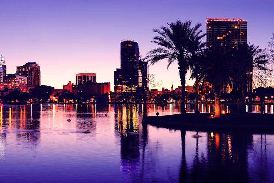 A city skyline at dusk with illuminated buildings reflecting on a calm body of water.