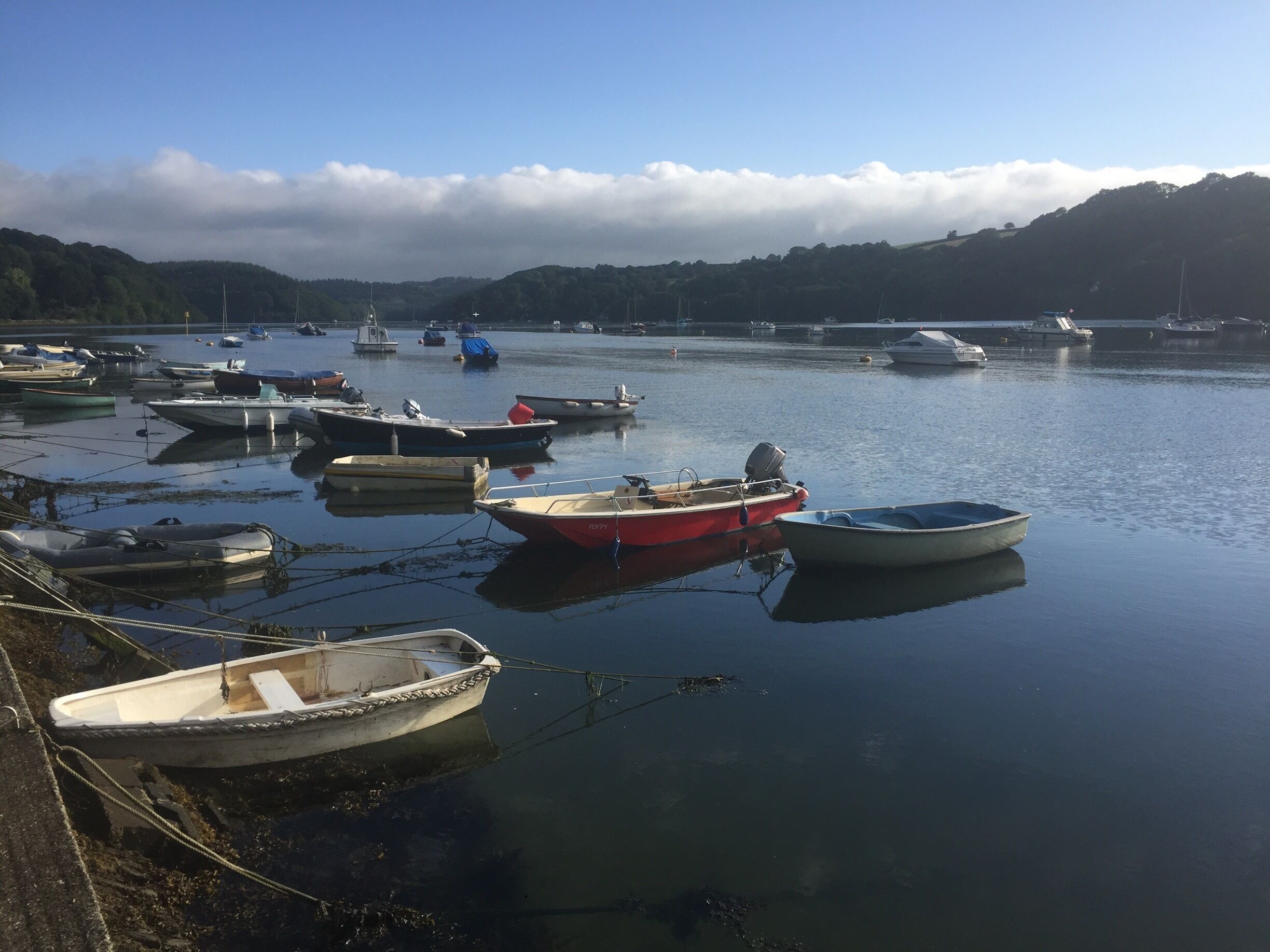 A calm harbor with several boats docked, surrounded by hills and a partly cloudy sky.