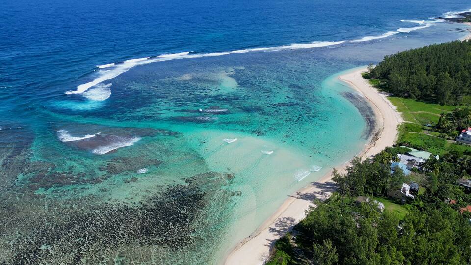 A coastal landscape with clear turquoise waters, a sandy beach, and a forested area with buildings.