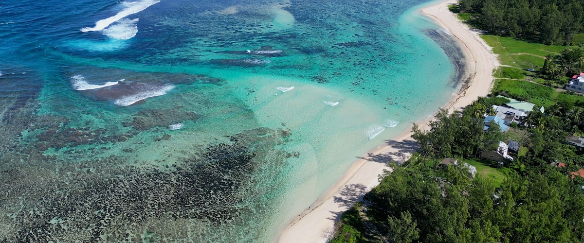 A coastal landscape with clear turquoise waters, a sandy beach, and a forested area with buildings.