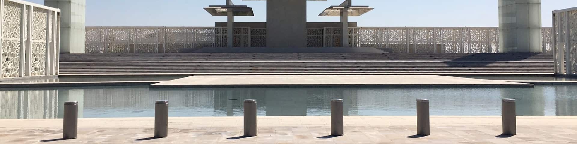 A modern architectural structure with a central table and two tall pillars, surrounded by a water feature and patterned fencing.
