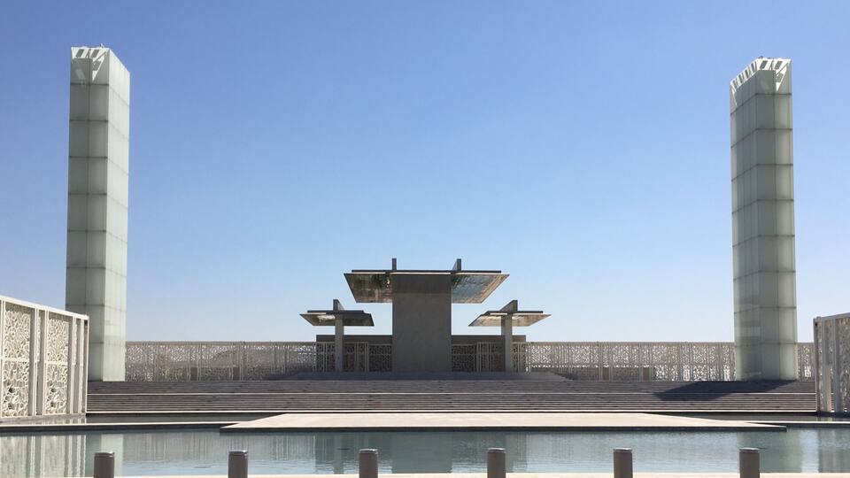 Ein modernes architektonisches Bauwerk mit einem zentralen Tisch und zwei hohen Säulen, umgeben von einem Wasserspiel und Zäunen mit Muster.