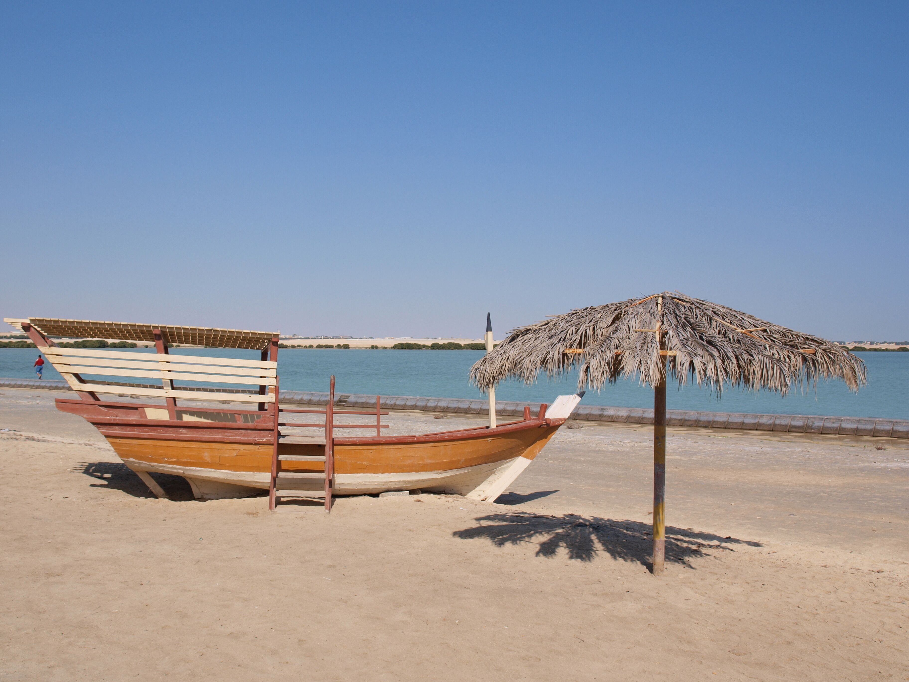 A wooden boat with a thatched roof on a sandy beach, a ladder, and a clear blue sky in the background.