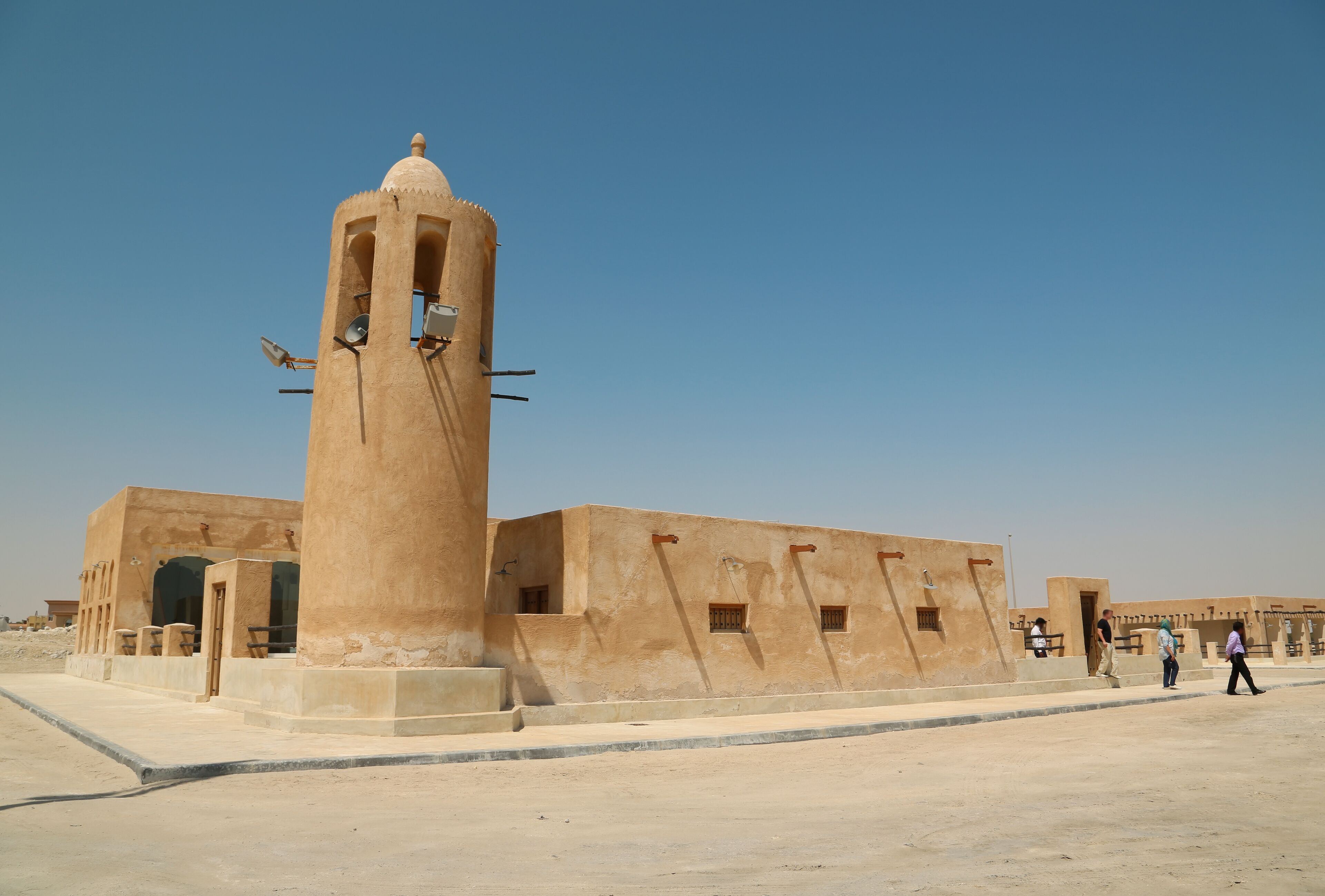 A large beige tower with satellite dishes, a building with arched doorways, and a clear blue sky.