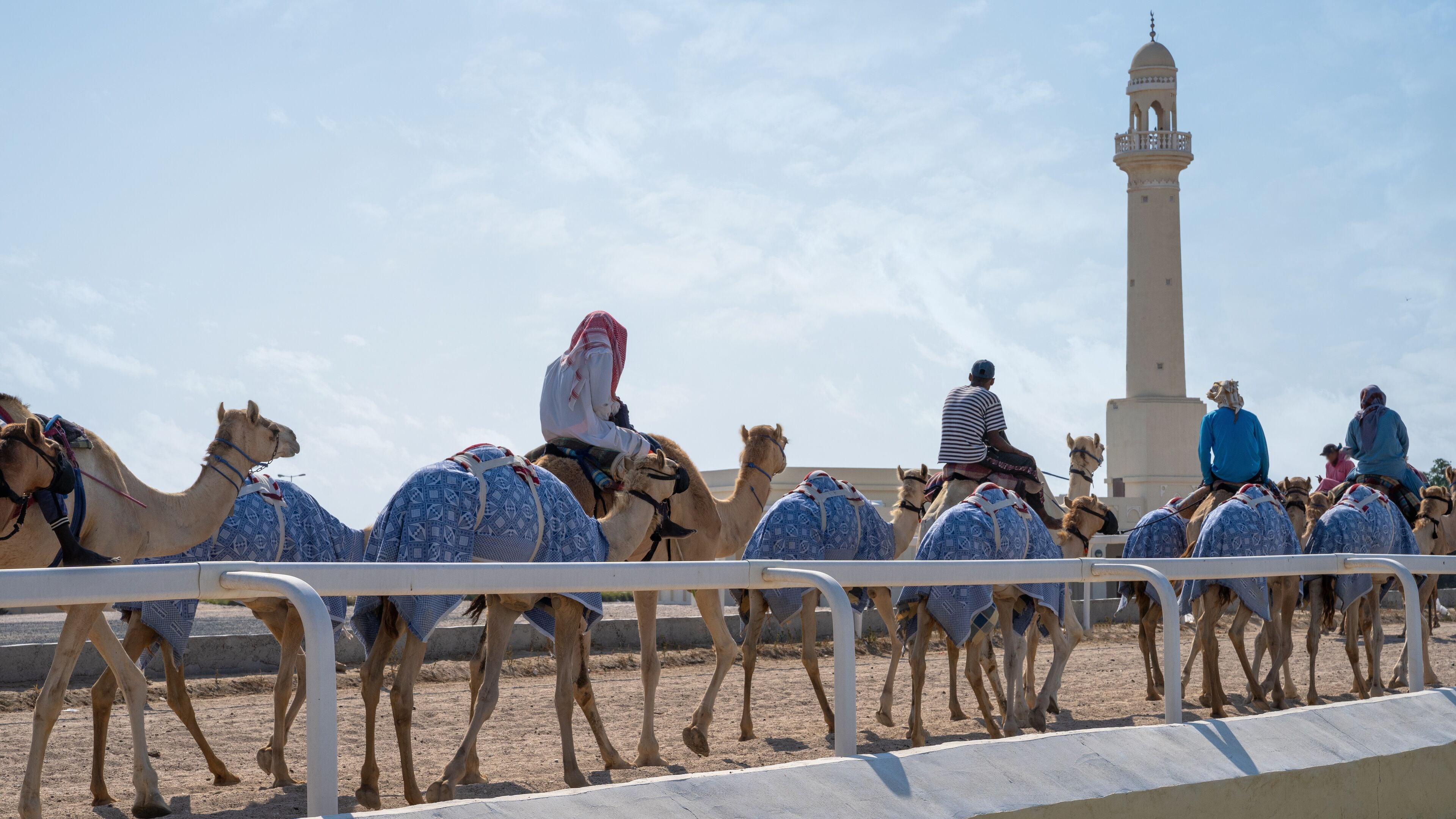 A group of camels with riders in traditional attire, a tall minaret, and a clear sky.