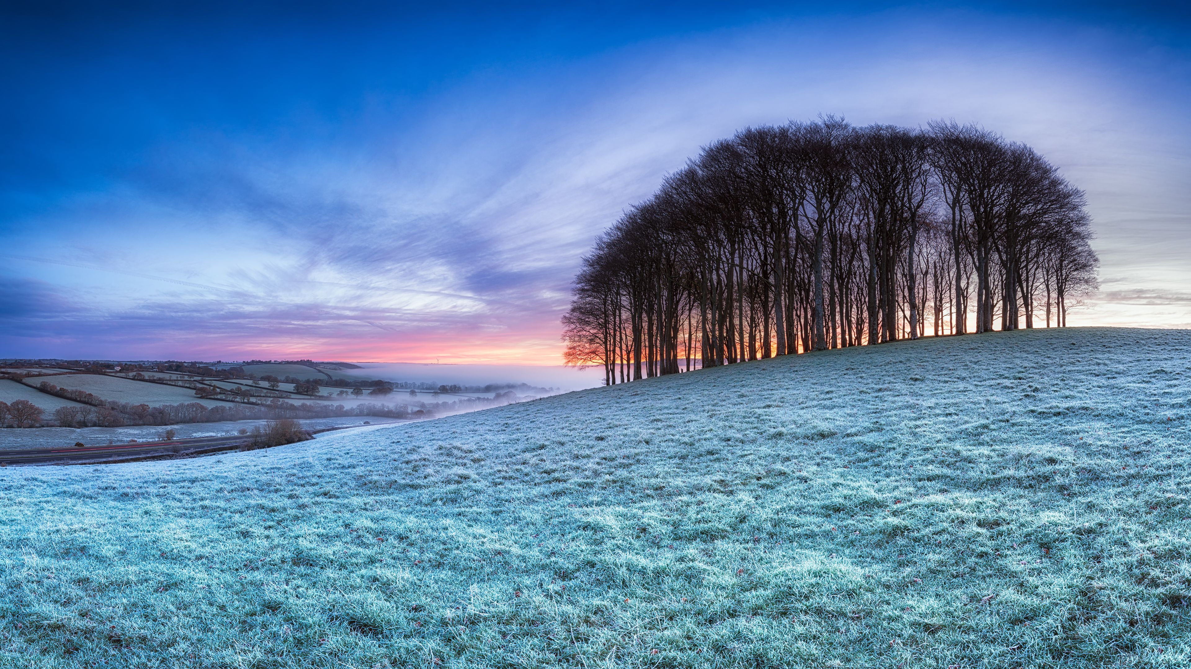A frost-covered landscape with a row of trees on a hill, a misty valley, and a sunset sky.